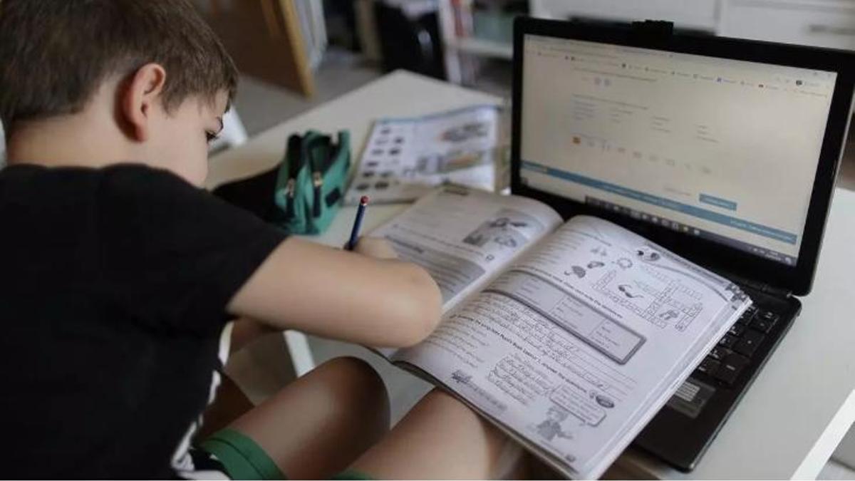 Niño pequeño escribiendo en un libro con un ordenador delante