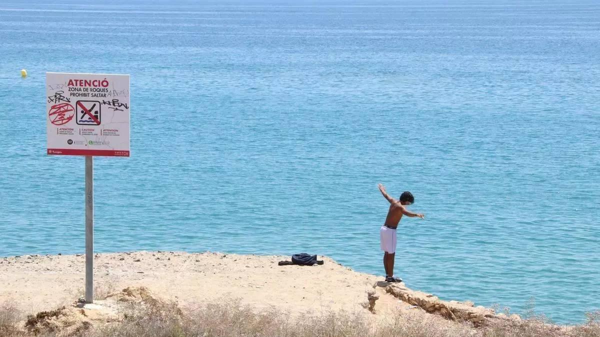 Un menor es disposa a saltar des de les roques de la platja del Miracle, a Tarragona, davant d’un cartell de prohibició