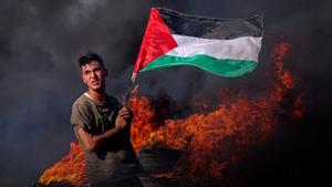 Un joven ondea la bandera de Palestina mientras arden a su lado neumáticos en una dura protesta junto a la valla fronteriza al este de la ciudad de Gaza