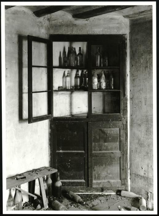 Fotografia d'un racó de la cuina d'una masia abandonada, hi trobem un armari encastat construït aprofitant una cantonada de la estança, una banqueta i ampolles de vidre buides a l'armari i escampades per terra