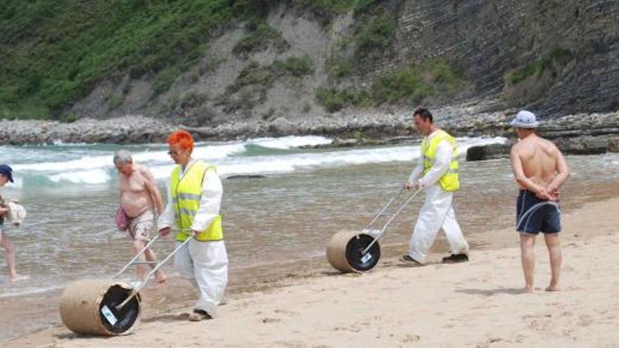 Dos operarios de limpieza pasan los rodillos por el arenal de Rodiles mientras varios bañistas pasean por la zona de la playa cerrada al baño.