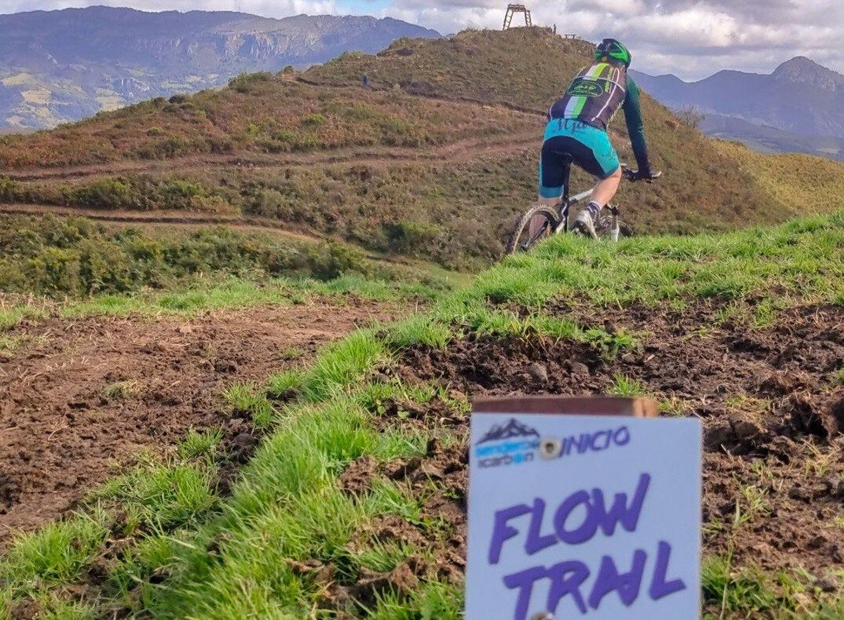 Un ciclista desciende por el circuito "flow trail" de Laviana, con el cuadro de mina que sirve de inicio al recorrido al fondo de la imagen. | Senderos del Carbón