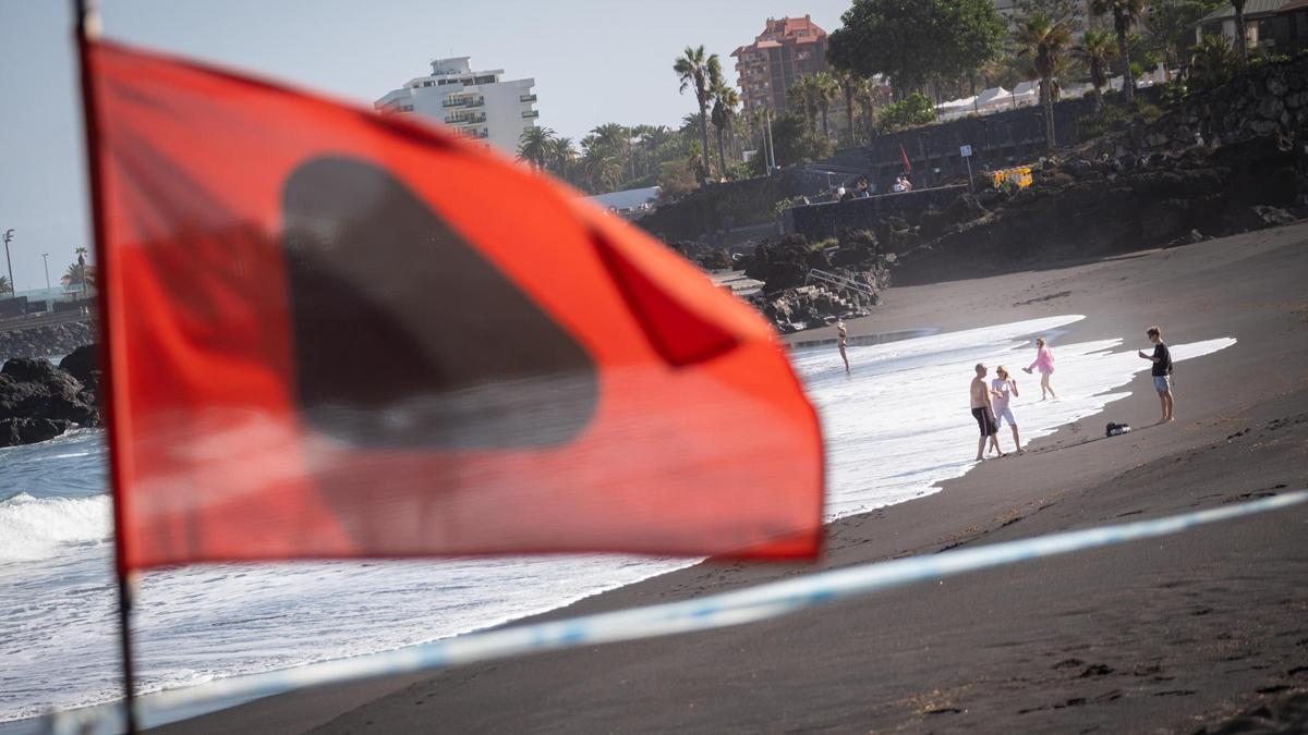 La bandera de prohibición de bañarse en Punta Brava y al fondo turistas remojándose en la orilla.