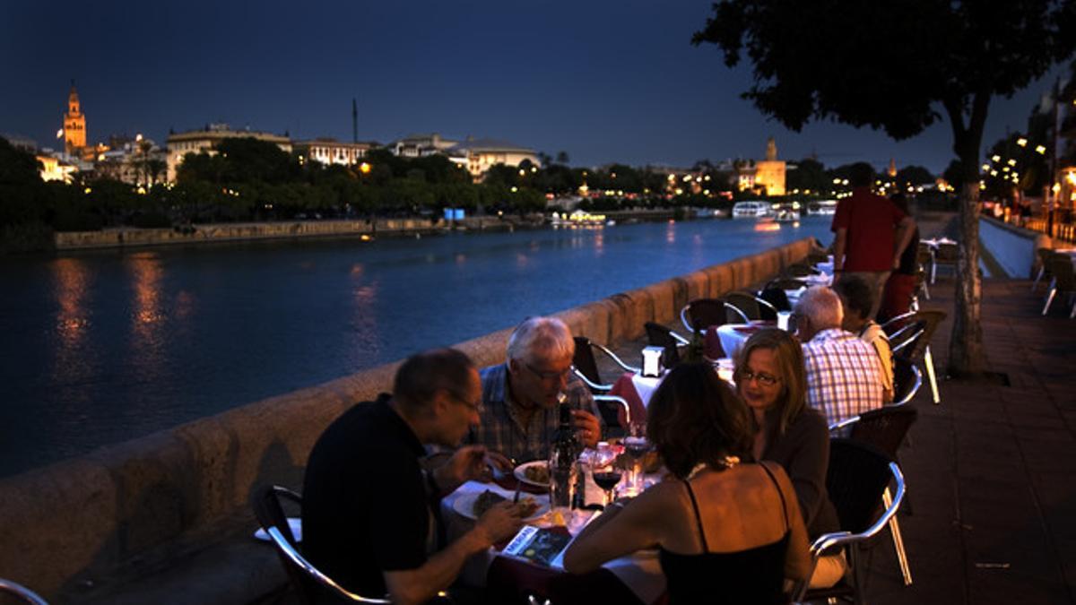 Unos turistas cenan en una terraza de la calle Betis de Sevilla, con vistas al río Guadalquivir y con la Giralda y la Torre del Oro al fondo.