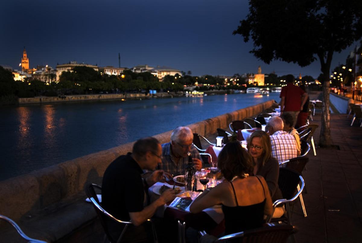 Uns turistes sopen en una terrassa del carrer Betis de Sevilla, amb vistes al riu Guadalquivir i amb la Giralda i la Torre del Oro al fons.