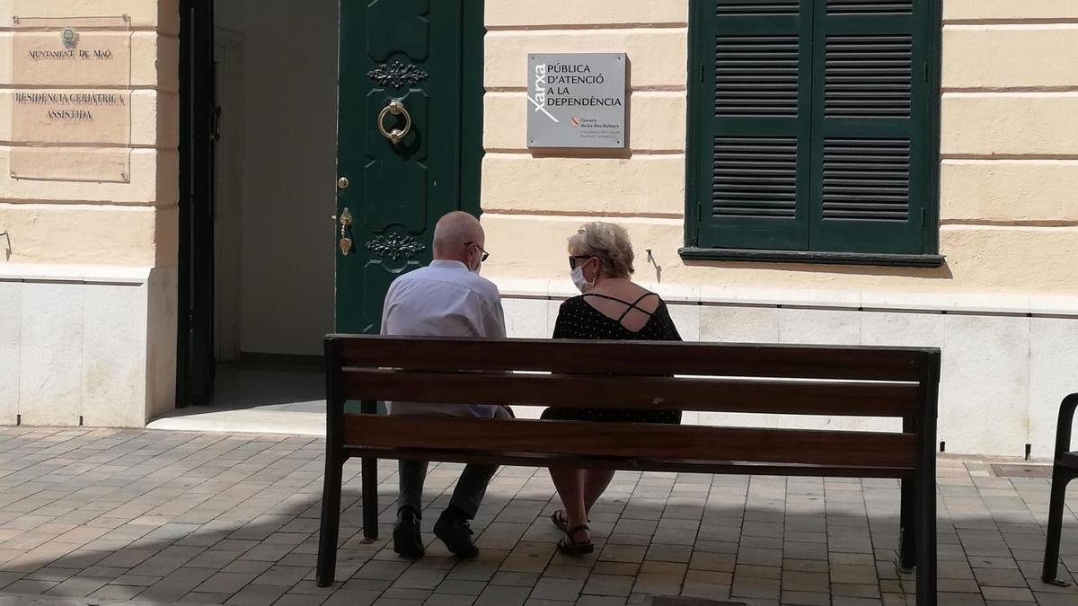 Dos personas mayores conversan sentadas en un banco y con mascarilla.