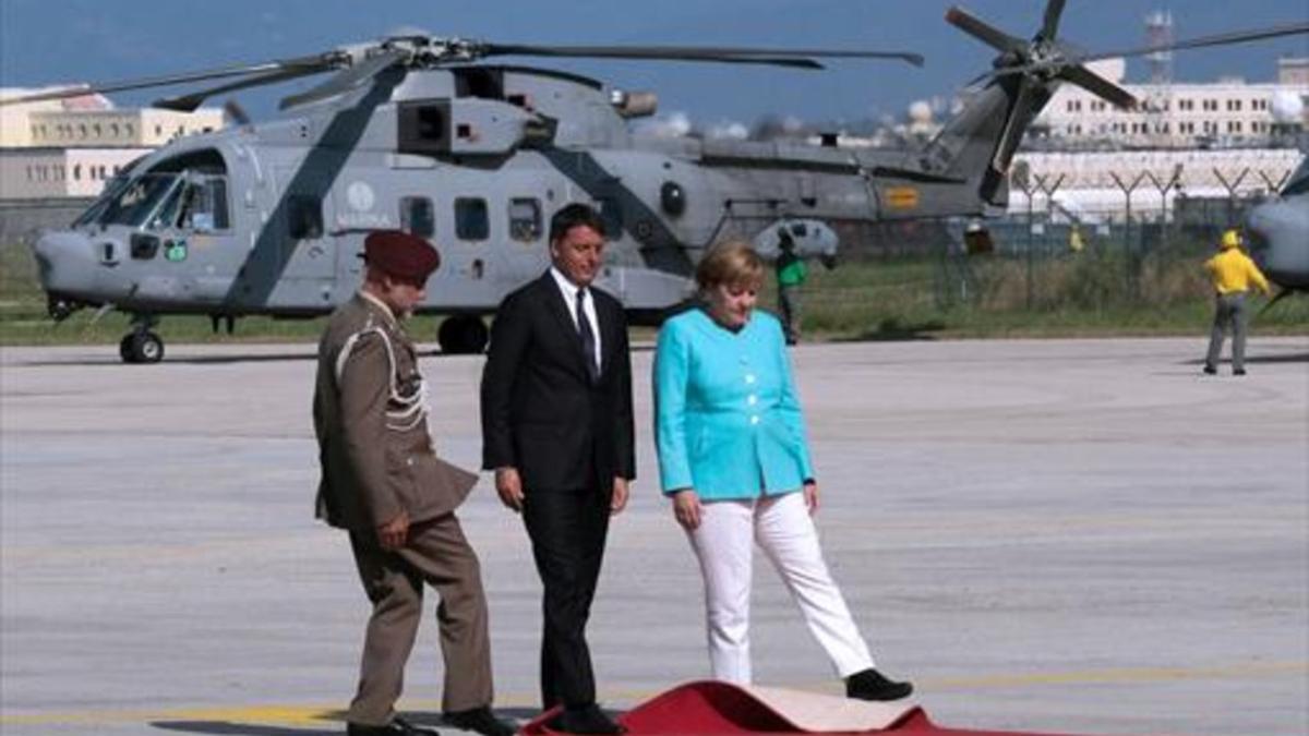 Viajera 8 Renzi, Merkel y un oficial tratan de ajustar la alfombra roja en el aeropuerto de Nápoles, antes de desplazarse a Ventotene.