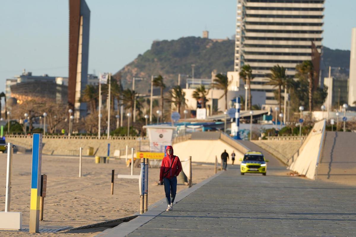 Una chica caminando por el paseo marítimo frente a la playa del Bogatell, en Barcelona, con un coche patrulla a su espalda.