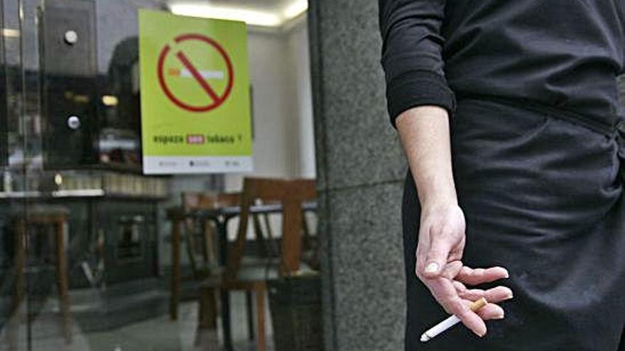 Una persona sostiene un pitillo, junto a un cartel de prohibido fumar en la cristalera de un bar.