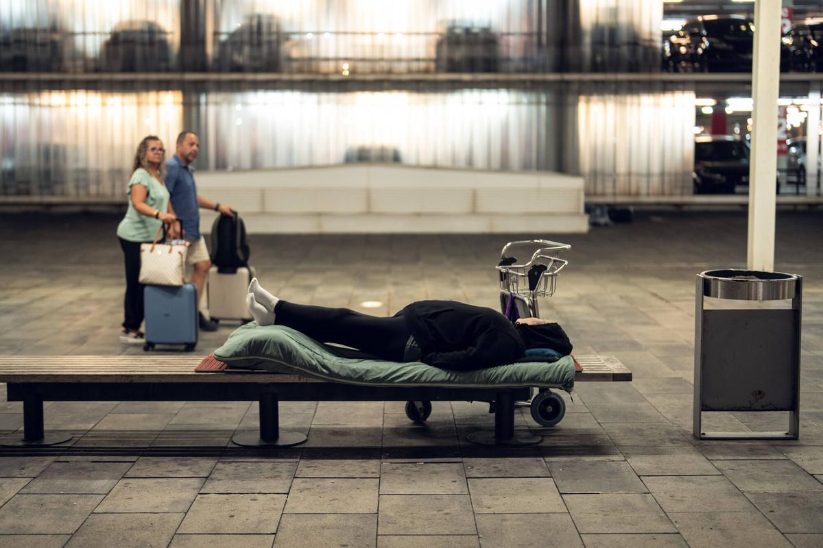 Dos viajeros observan una persona sin hogar que duerme en un banco en los exteriores de la terminal T1 del aeropuerto de Barcelona.