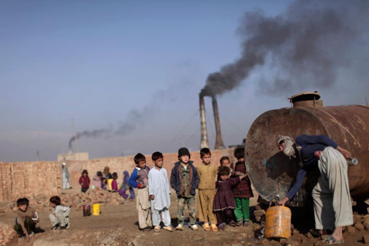 Un grup de nens afganesos observen com un home omple un bidó d’aigua en un camió cisterna. Al fons, la silueta d’unes fàbriques de totxo, als afores de Kabul (Afganistan).