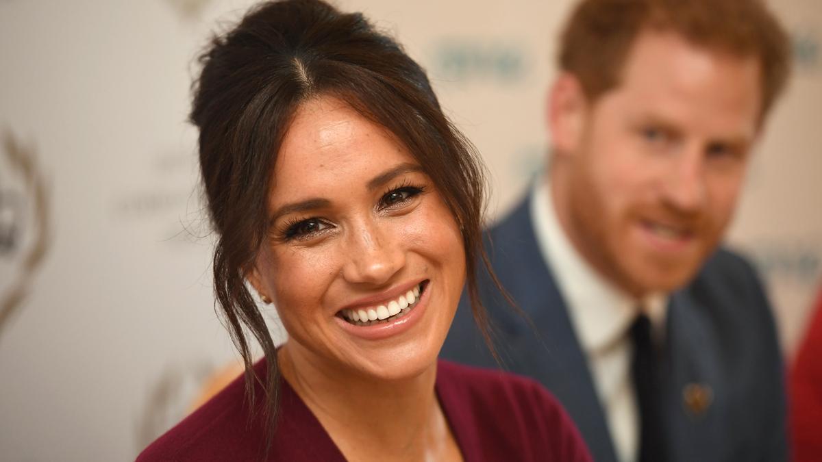 Primer plano de Meghan Markle con el principe Harry de fondo