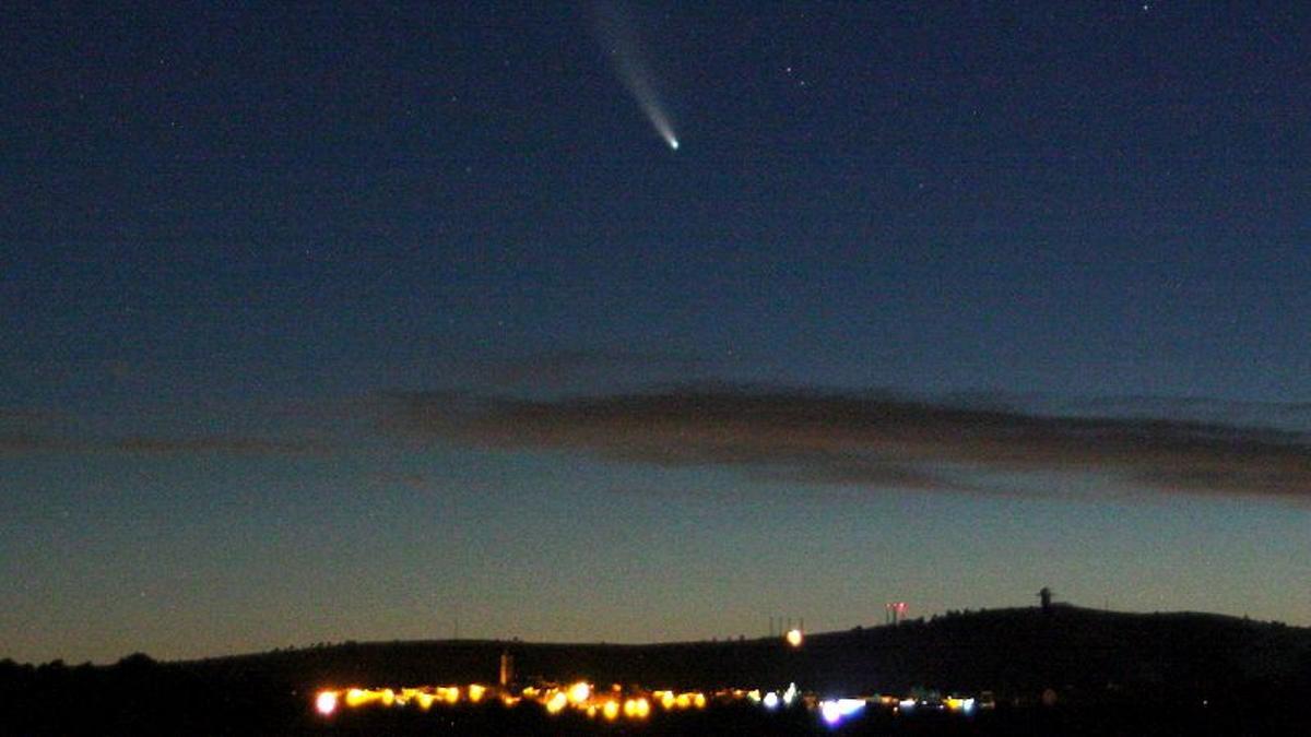 El cometa Neowise, fotografiado encima de un centro habitado fuertemente iluminado.