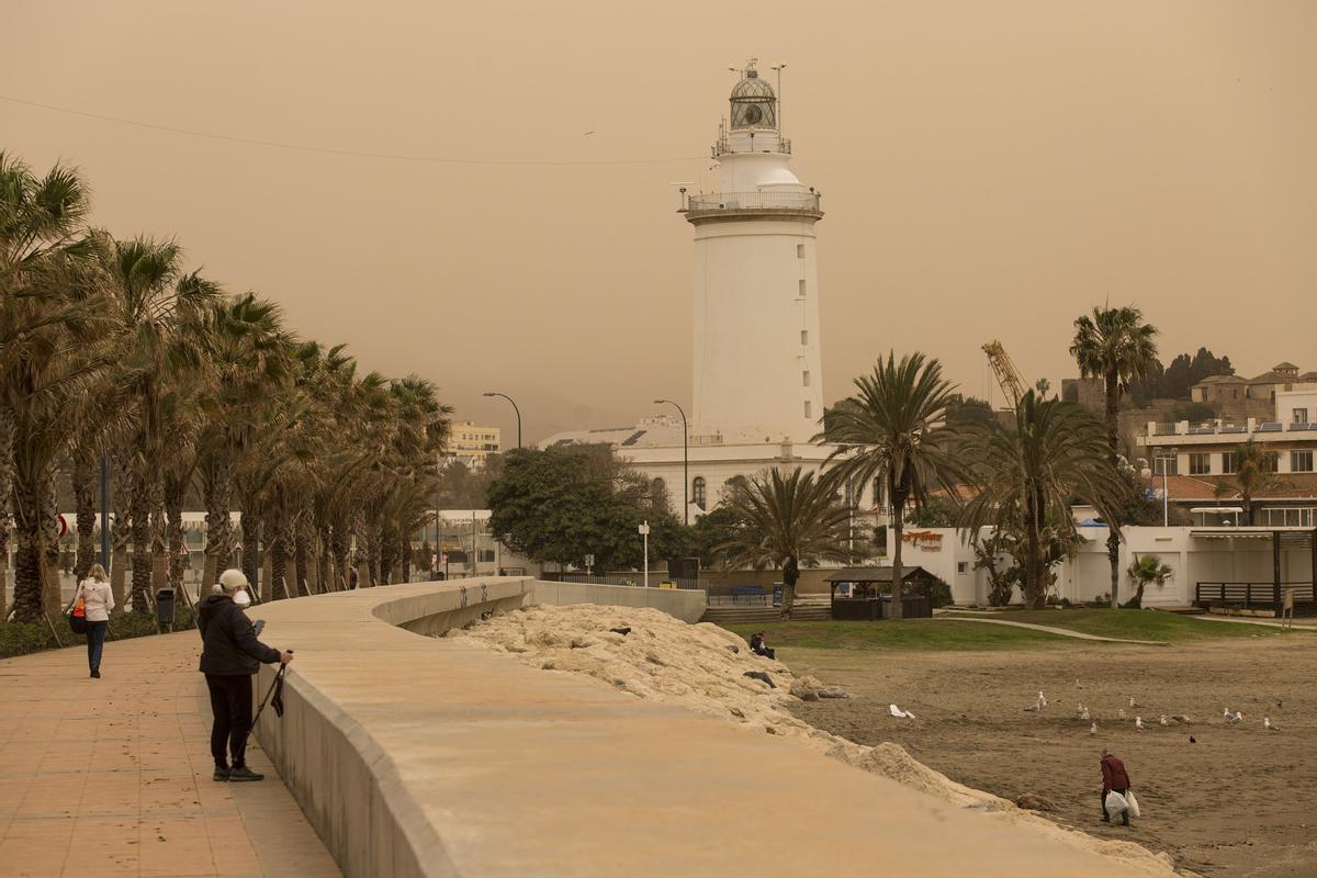 Varias personas caminan por la zona del Puerto de Málaga, con La Farola al fondo, cubierta por polvo procedente del desierto del Sáhara.