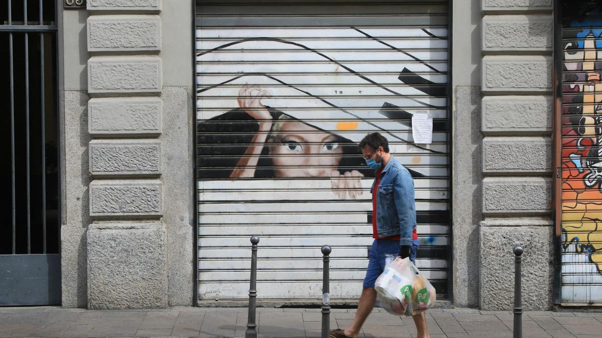 Un ciudadano camina frente a un local comercial con la persiana bajada.