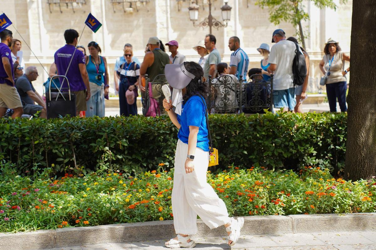 Turista caminando por el centro de Sevilla durante un día de calor, equipada con un sombrero de ala ancha y un ventilador portátil de mano para refrescarse mientras