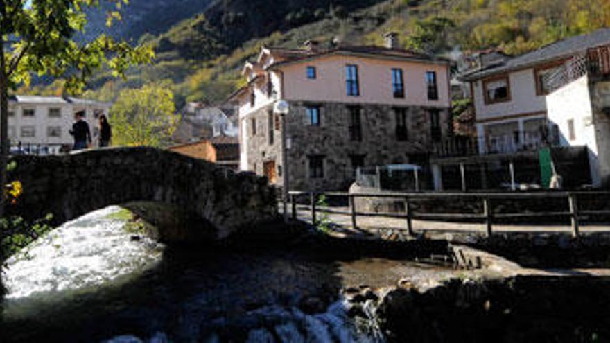 Dos personas cruzan el puente viejo de Pola de Somiedo, con Peñamellera detrás.