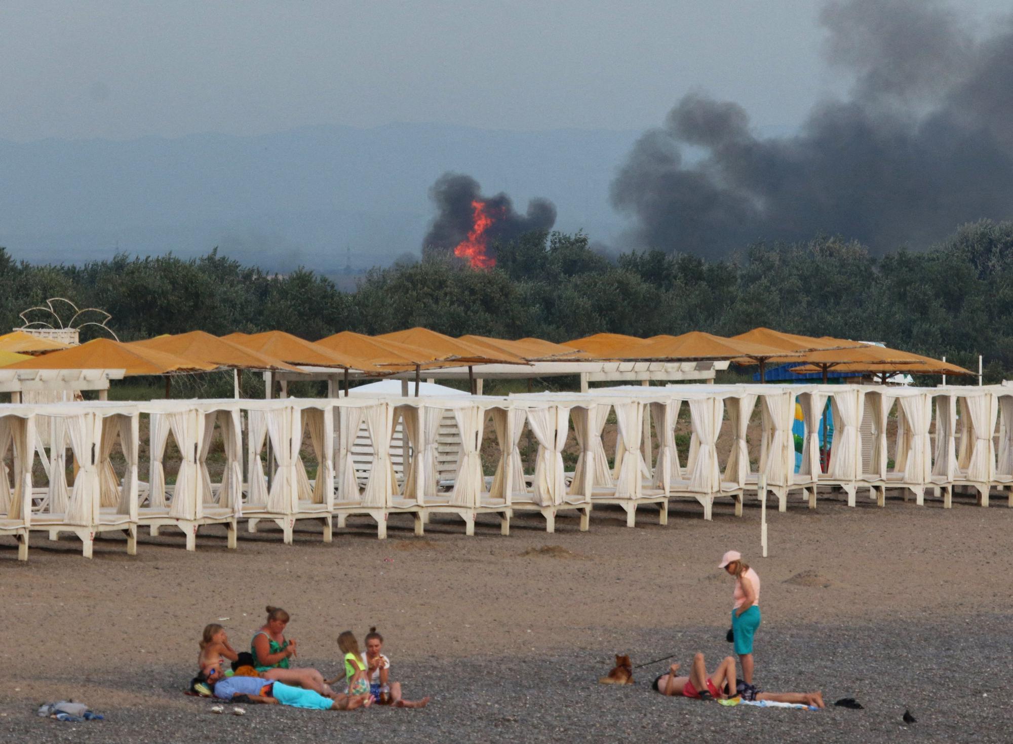 People rest on a beach as smoke and flames rise after explosions at a Russian military airbase, in Novofedorivka, Crimea August 9, 2022.