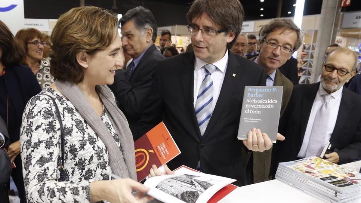 Carles Puigdemont le muestra a Ada Colau, durante la inauguración de Liber, el libro 'Si els alcaldes governessin el món'