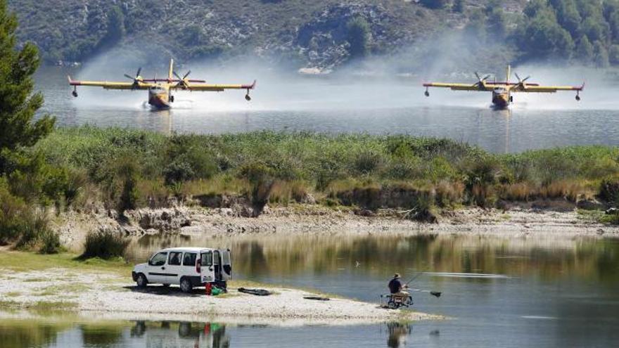 Dos hidroaviones amerizan en el embalse de Beniarrés para recoger agua, mientras un pescador observa la maniobra.