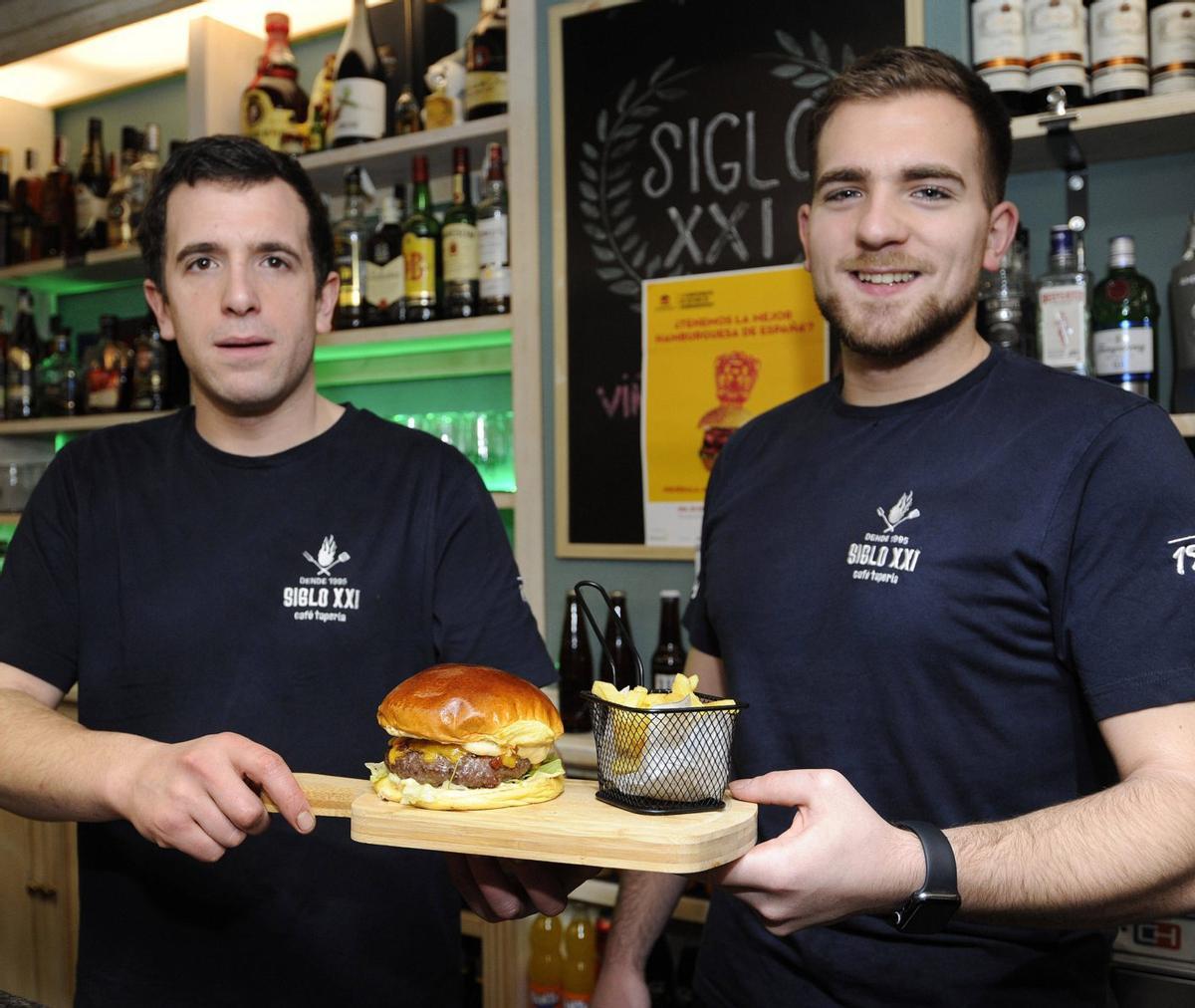 Alejandro y Diego Gil sostienen un ejemplar de la hamburguesa «La Nº 23» en la Tapería Siglo XXI. | BERNABÉ/JAVIER LALÍN