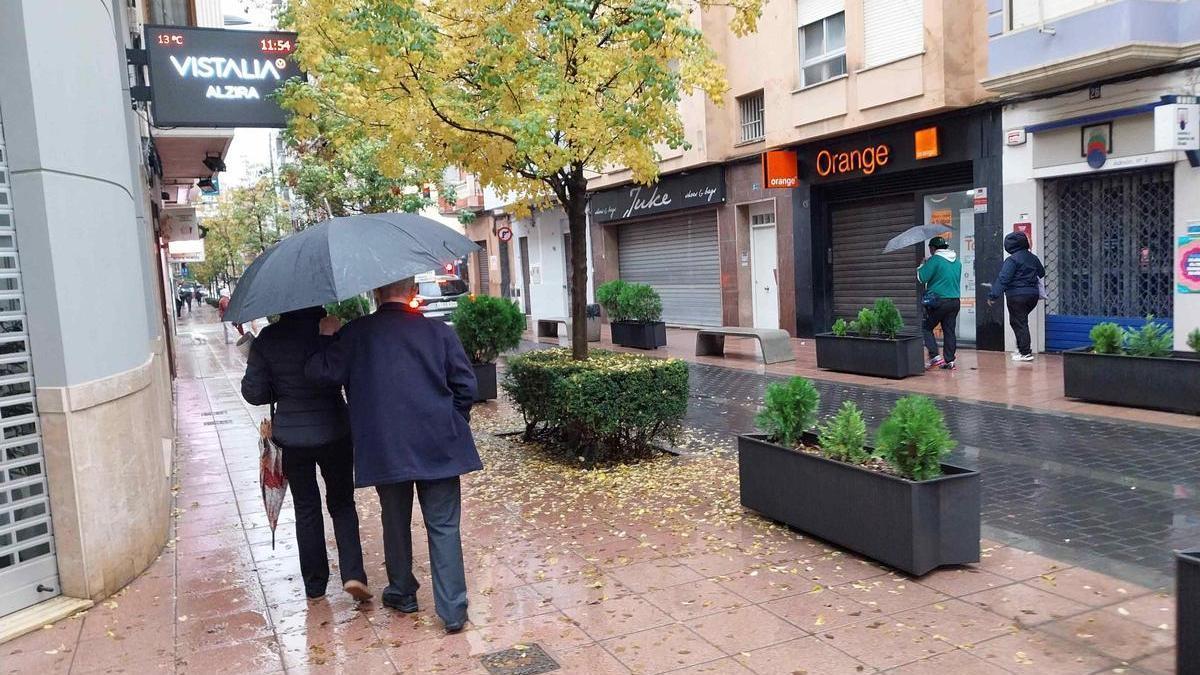 Varios peatones se protege con paraguas de la lluvia, este domingo, en la calle Pérez Galdós de Alzira.