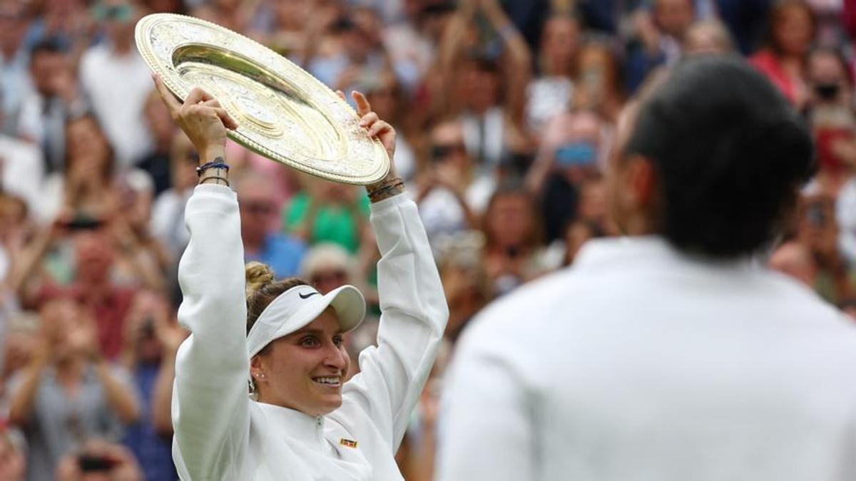 Vondrousova muestra el trofeo de campeona de Wimbledon, con Jabeur, de espaldas, mirándola.