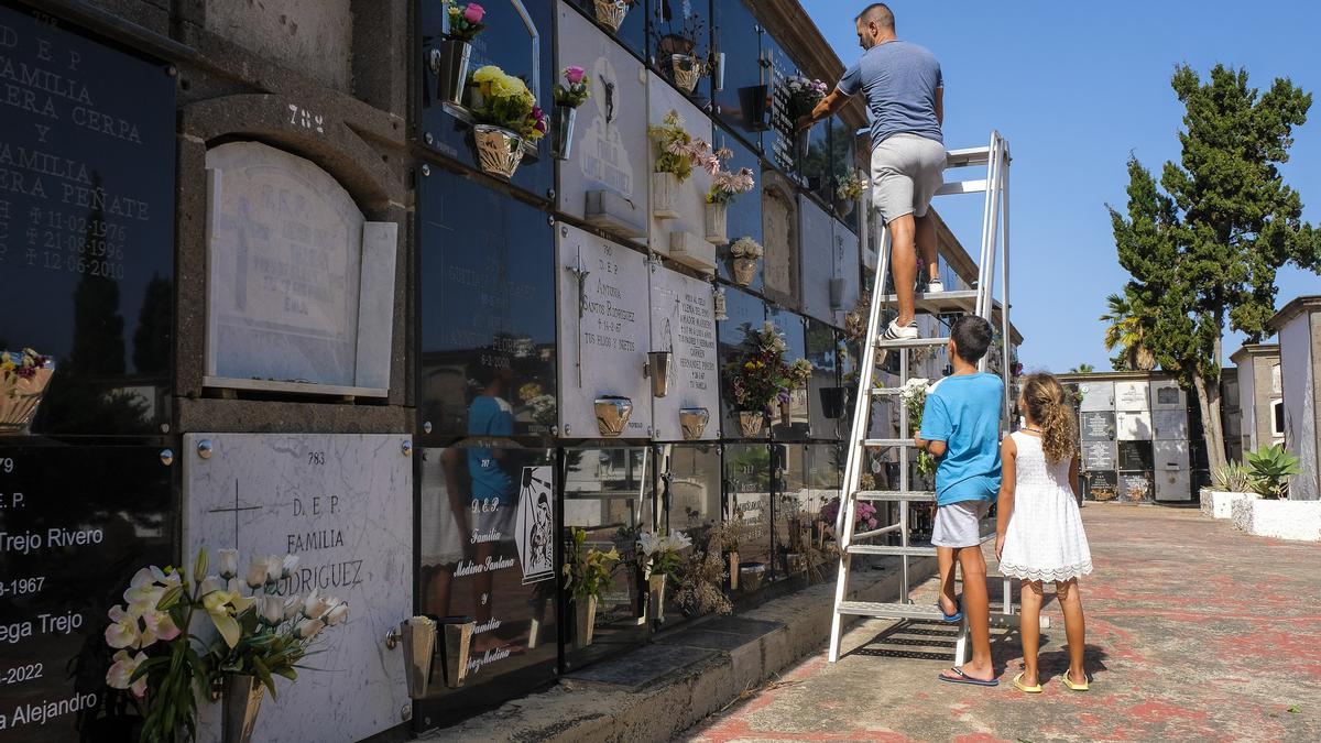 Un padre subido a una escalera coloca flores en un nicho en el cementerio de San Lázaro bajo la atenta mirada de sus hijos 