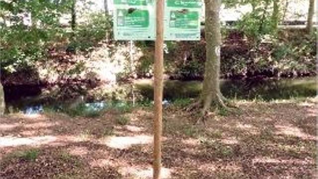 El cartell en primer pla i el curs del riu Fluvià al seu darrera.
