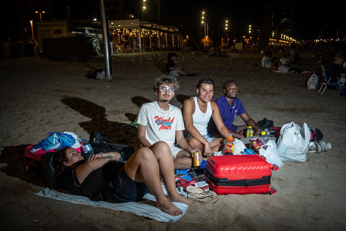 De izquierda a derecha, Miguel, de 19 años; Daniel, de 20; e Ismael, de 21, durante la entrevista. Los tres jóvenes, residentes en Londres, pasarán la noche en la playa de Barcelona para evitar el coste del alojamiento antes de continuar su viaje. A 6 de agosto de 2026. Sociedad. AUTOR: MANU MITRU