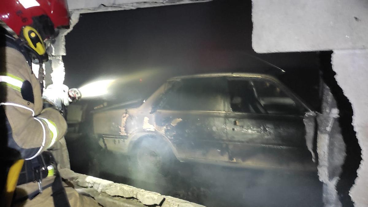 Un bombero rompe un muro junto a un coche quemado.