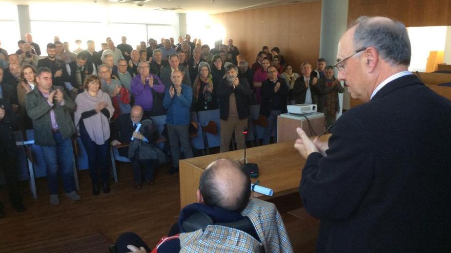 Tino Guimeráns, de espaldas en la silla de ruedas, con José Ramón Muñiz a su lado y el salón de plenos en pie al final de su intervención. // Gonzalo Núñez