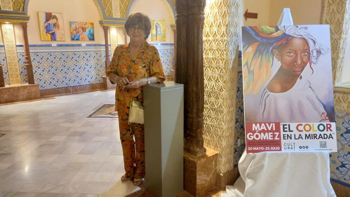 Mavi Gómez posa con el cartel de la exposición y, de fondo, algunas de sus coloridas obras pictóricas