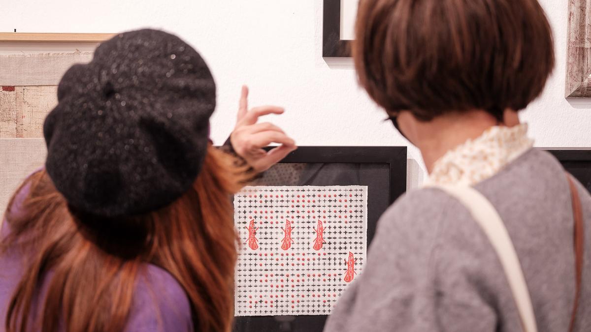 Dos mujeres observan varias obras de esta muestra colectiva.