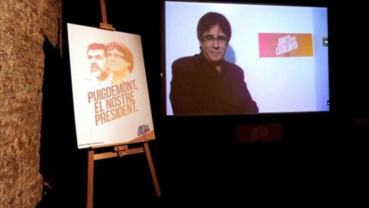 Videoconferencia de Puigdemont, junto a un cartel de Junts per Catalunya.