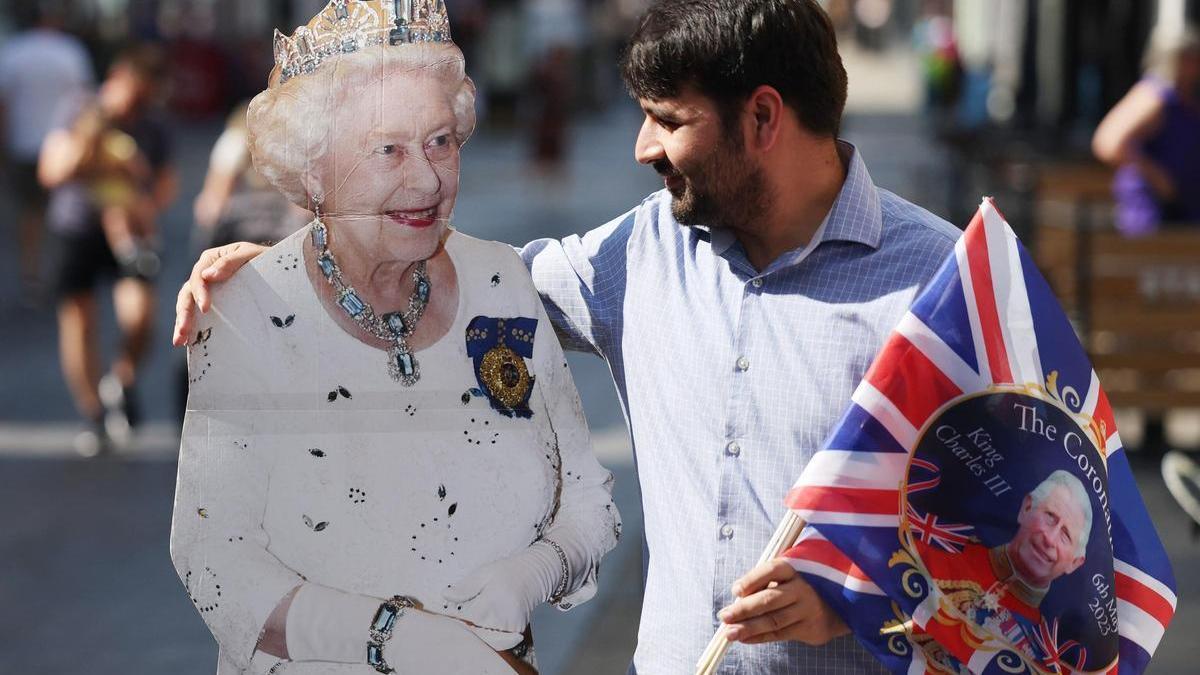 Un hombre posa con una fotografía a tamaño real de Isabel II y unos banderines de la coronación de Carlos III, este jueves en Windsor. /