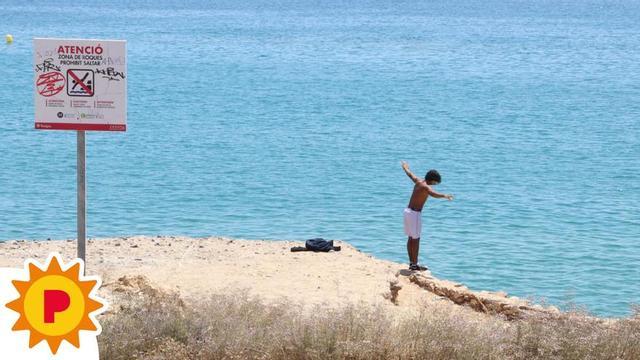 Un menor procedeix a saltar des de les roques de la platja del Miracle a Tarragona, davant un cartell de prohibició.