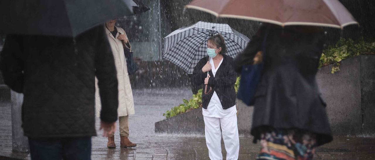 Una mujer espera para cruzar resguardándose de la lluvia debajo de un paraguas y ataviada con una mascarilla.