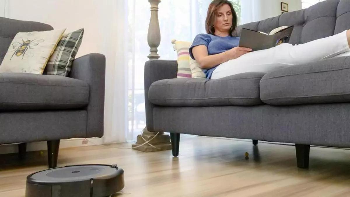 Un robot aspirador limpia el suelo mientras una mujer lee