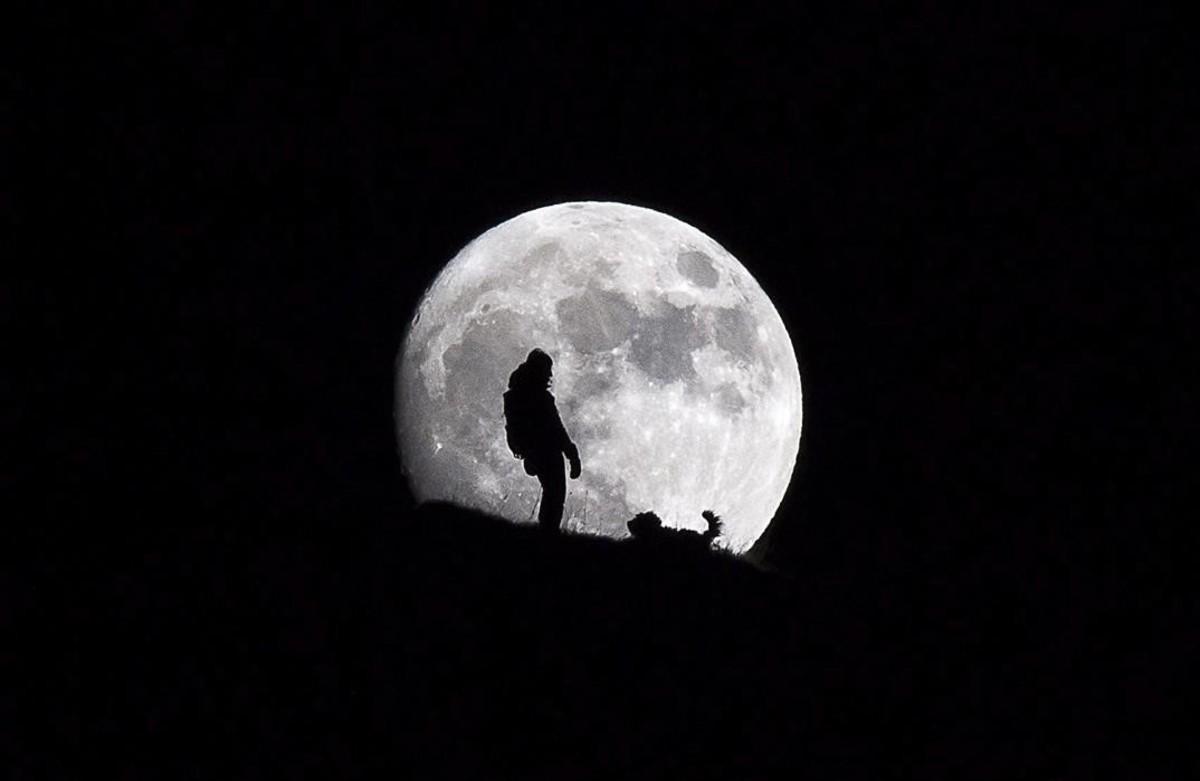 Silueta d’una dona mentre passeja el gos amb la lluna plena com a teló de fons al cim de Croix des Chaux, als Alps suïssos.