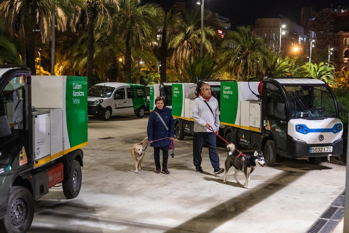 Dos vecinos pasean a sus perros entre vehículos de limpieza con el lema del consistorio ’Cuidem Barcelona’.