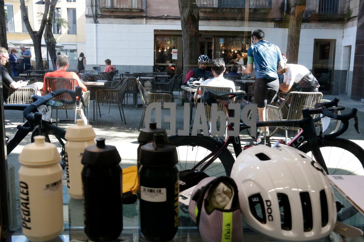 En primer terme, cascos i bidonets i, al fons, uns ciclistes en una terrassa de Girona