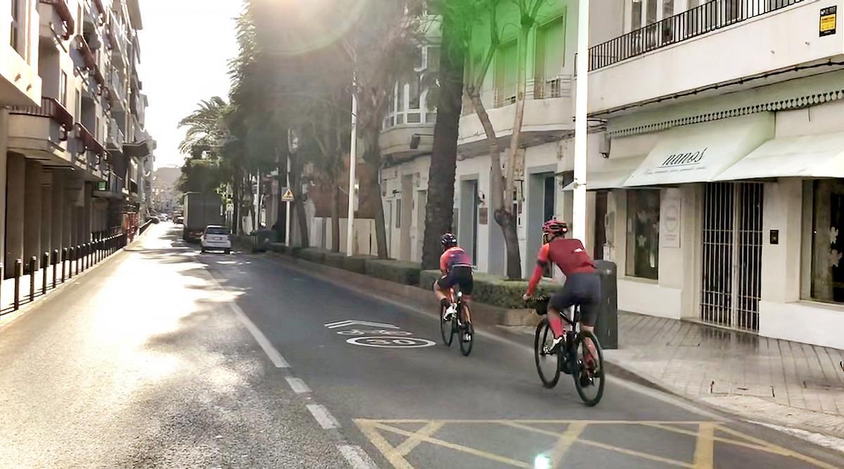 Ciclistas circulan por travesía de la N332 en el casco urbano de Altea junto a las señales horizontales de límite de velocidad.