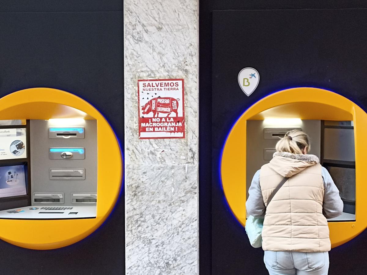 Una vecina de Bailén saca dinero de un cajero en el centro del pueblo, junto a un cartel contra la macrogranja.