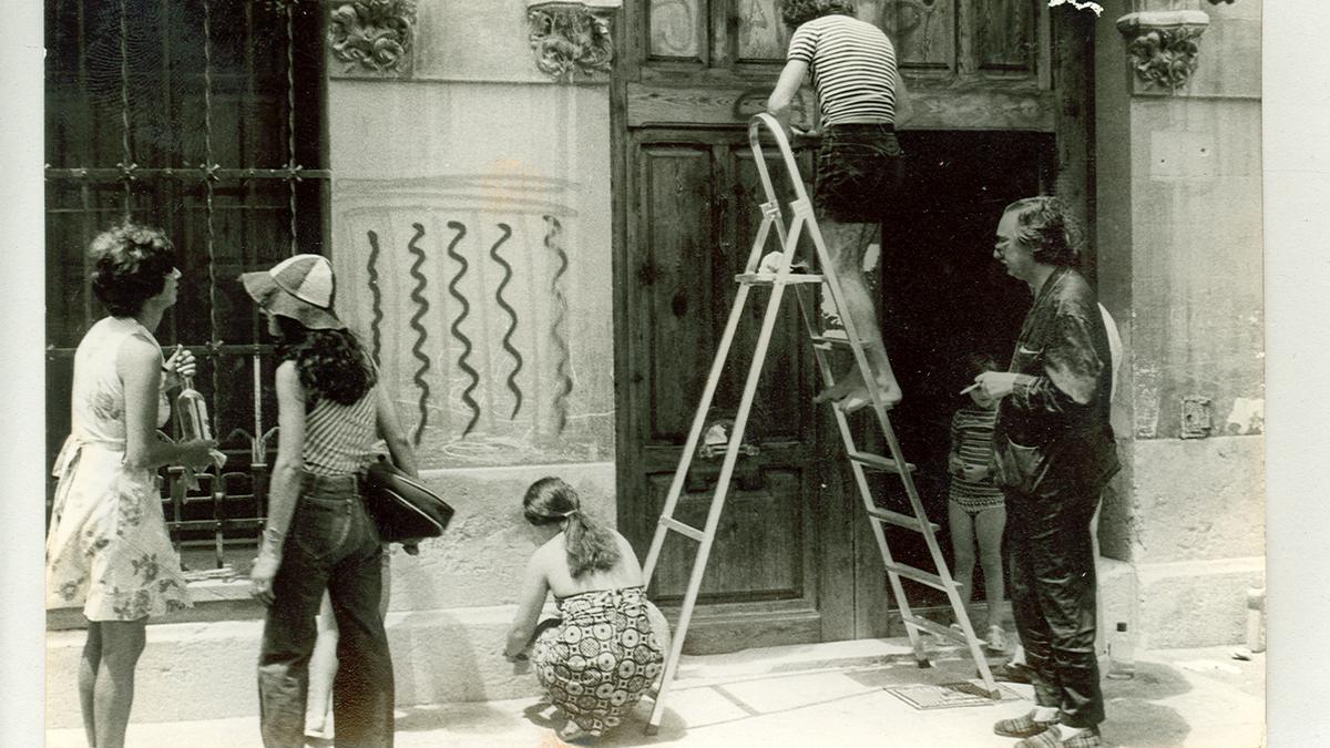 Joan Fuster, a la dreta de la imatge, observa com diverses persones netegen les pintades fetes a sa casa pel dia de Sant Jaume. Autor desconegut. 1977.