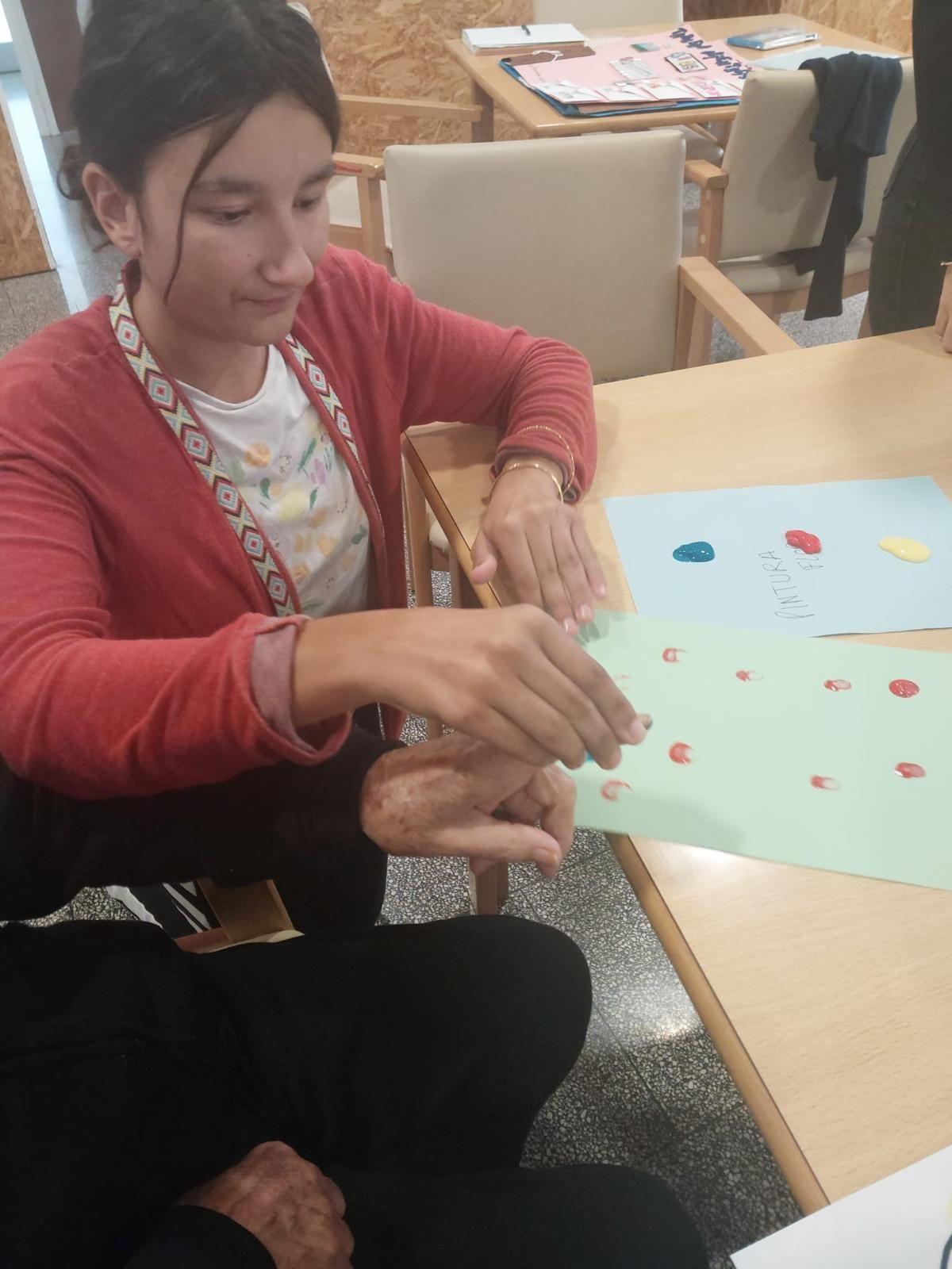 Una alumna ayuda a pintar con los dedos a una persona con dependencia.