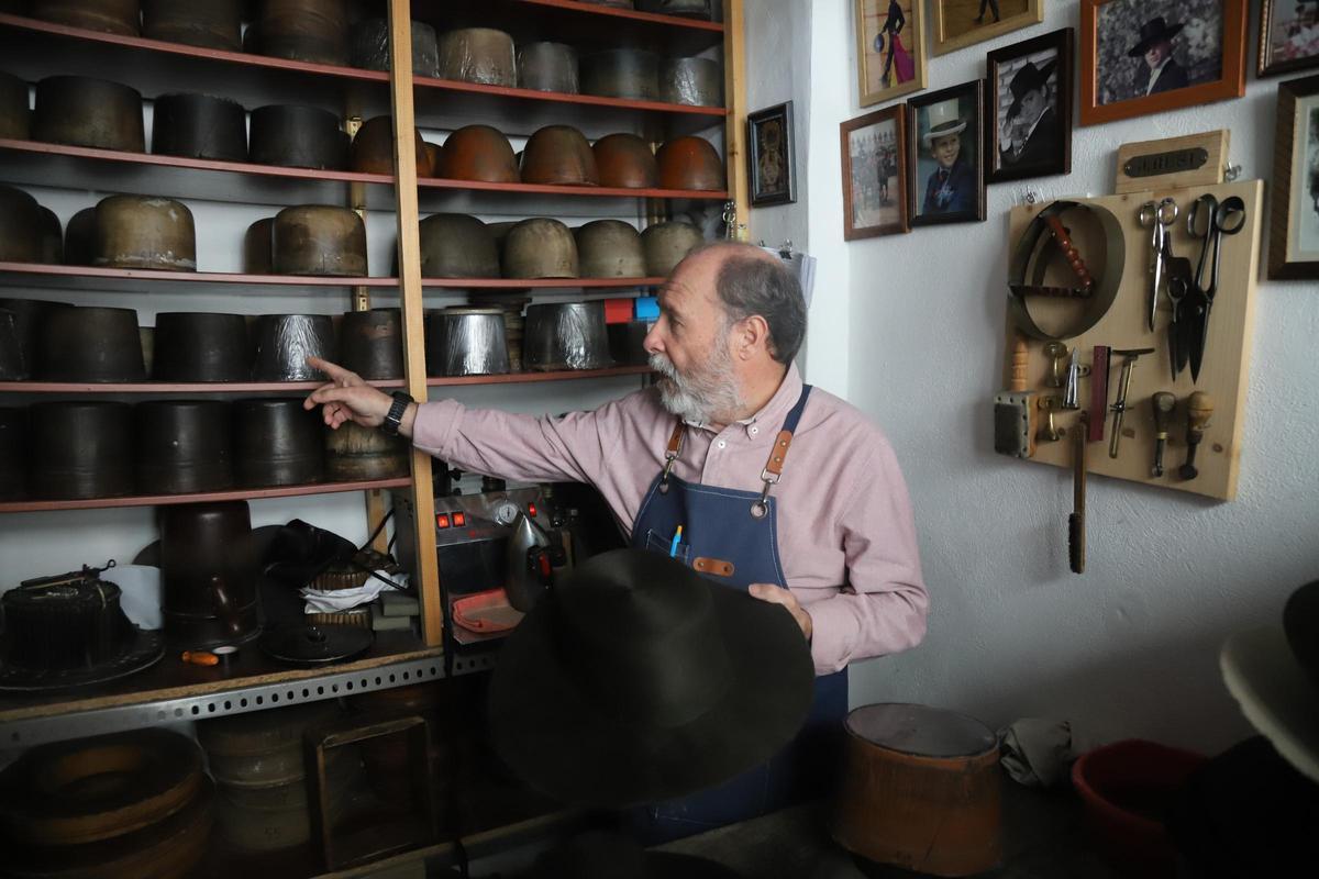 Mario Roldán sostiene un sombrero en el que trabaja, en su pequeño taller, con un estante de hormas y herramientas de fondo.