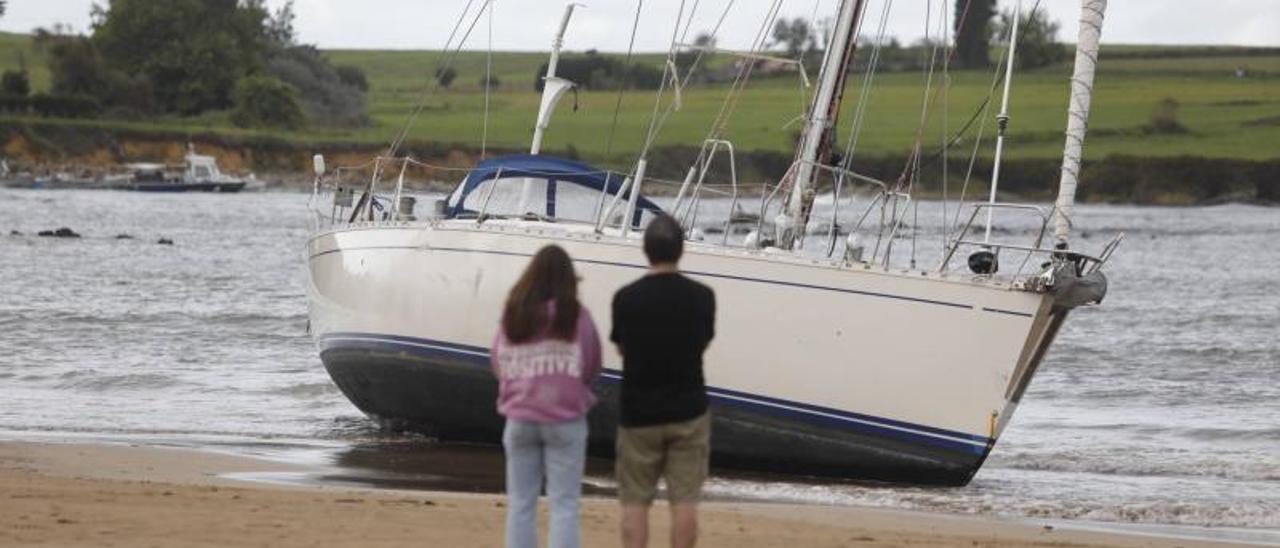 Dos personas contemplan desde la arena el velero varado en Bañugues. | R. Solís