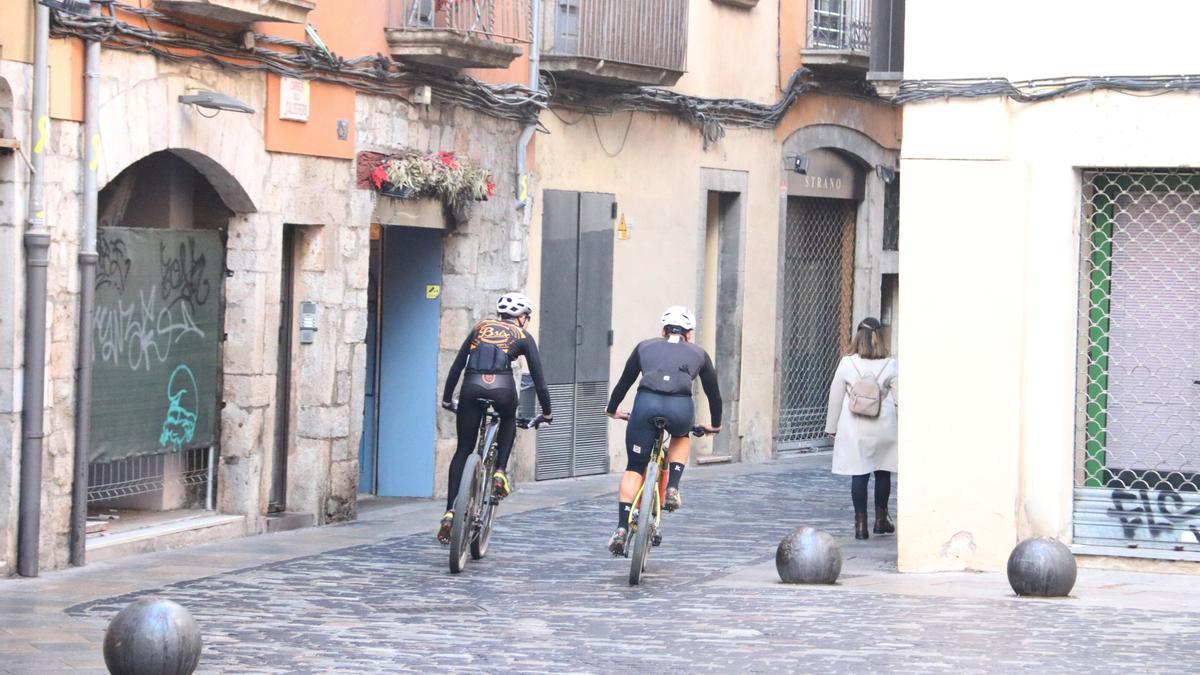 Dos ciclistes passen pel Barri Vell de Girona al costat d&#039;una vianant.