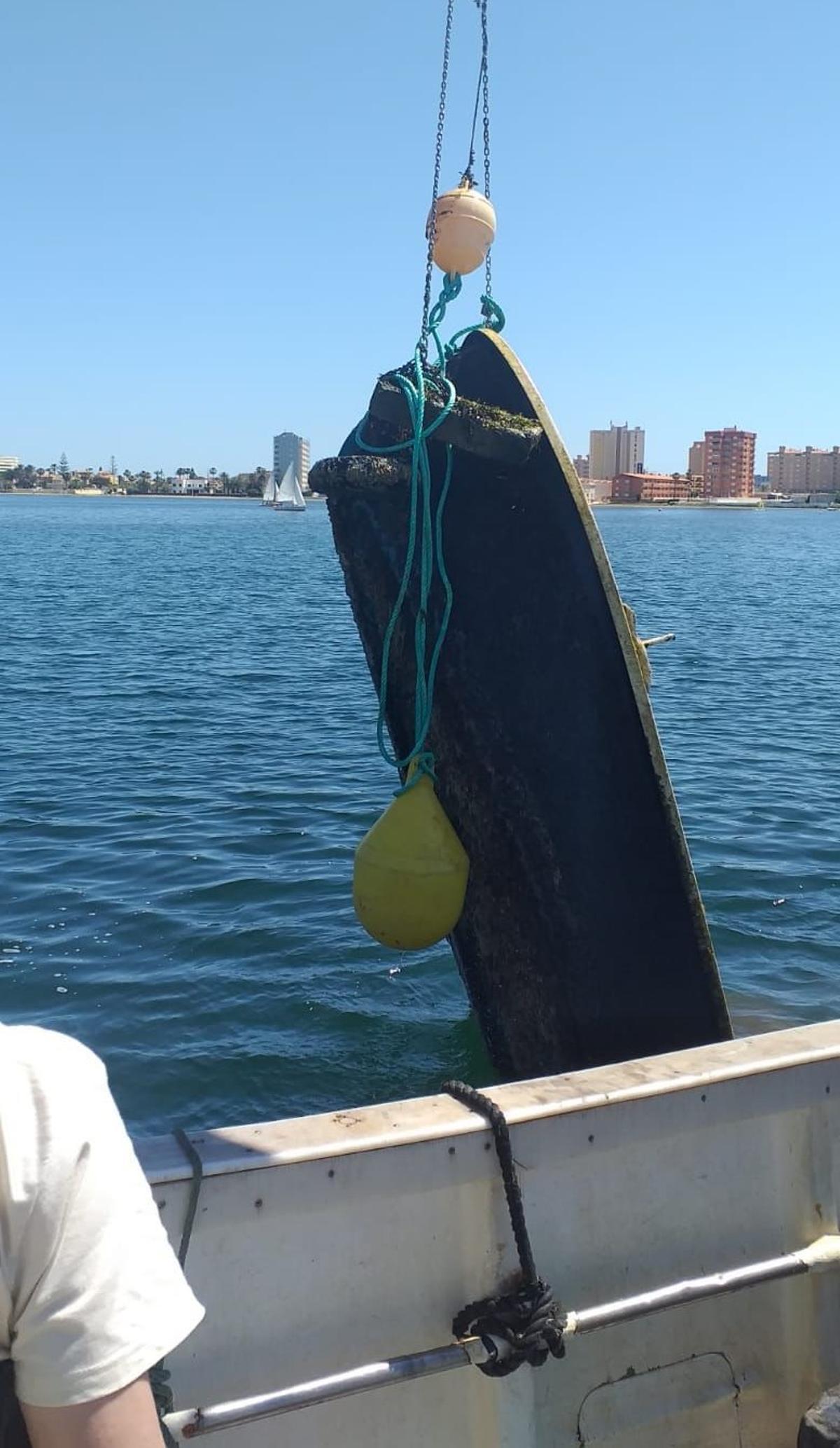 Un barco saca de las aguas del Mar Menor una embarcación abandonada.